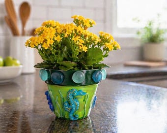 Può includere: Vaso di fiori dipinto a mano con una pianta fiorita gialla. Il vaso verde è decorato con motivi di cavallucci marini e alghe blu, e perline di vetro blu e trasparenti. Un oggetto decorativo per la casa, che aggiunge un tocco di colore.