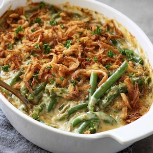 May include: A casserole dish filled with green bean casserole. The dish features green beans in a creamy sauce, topped with crispy fried onions and fresh parsley. The casserole is in a white oval baking dish.
