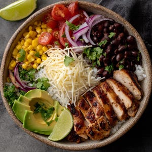 Puede incluir: Un cuenco colorido lleno de ingredientes frescos. Incluye pollo a la parrilla, rodajas de aguacate, maíz, tomates, cebolla roja, frijoles negros, queso rallado, cilantro y una rodaja de lima. El cuenco está sobre una superficie gris.