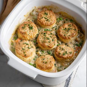May include: A white casserole dish filled with chicken pot pie. The pot pie has a creamy sauce with visible peas and carrots, topped with six golden-brown biscuits sprinkled with fresh parsley. The dish is set on a white surface.