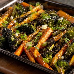 Op de afbeelding: Een close-up van geroosterde groenten in een bakplaat. Het gerecht bevat feloranje wortelen en groene broccoliroosjes, bestrooid met sesamzaad en gehakte groene uien. De groenten zijn gerangschikt in een rechthoekige metalen bakplaat.