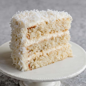 Peut inclure: Une part de gâteau à la noix de coco sur un présentoir à gâteau blanc. Le gâteau a plusieurs couches avec du glaçage blanc et est recouvert de noix de coco râpée. Le gâteau est de couleur beige clair et repose sur une assiette blanche.