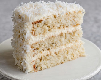 May include: A slice of coconut cake on a white pedestal cake stand. The cake has multiple layers with white frosting and is covered in shredded coconut. The cake is a light beige color and sits on a white plate.