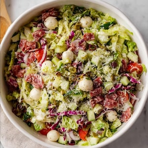 May include: A fresh salad in a white bowl, featuring a mix of green lettuce, red onion, cucumber, tomatoes, salami, mozzarella balls, and grated cheese. The salad is a vibrant mix of colors and textures, perfect for a light meal.