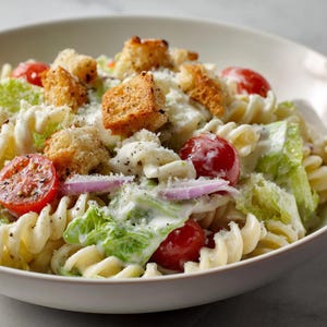 Puede incluir: Una ensalada César en un cuenco blanco. La ensalada incluye pasta rotini, picatostes, tomates cherry, cebolla roja y lechuga romana, todo cubierto con un aderezo cremoso y espolvoreado con queso.