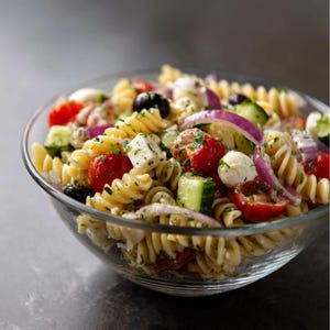 Könnte beinhalten: Eine Glasschüssel mit Nudelsalat. Der Salat enthält Rotini-Nudeln, rote Kirschtomaten, schwarze Oliven, rote Zwiebelscheiben, Gurkenstücke und weiße Käsewürfel. Der Salat ist mit einem leichten Dressing angemacht.