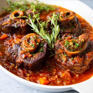 May include: A close-up shot of a white oval dish filled with braised meat shanks in a rich, red sauce. Fresh herbs, including rosemary and thyme, garnish the dish. The meat is tender, and the sauce has visible vegetables.