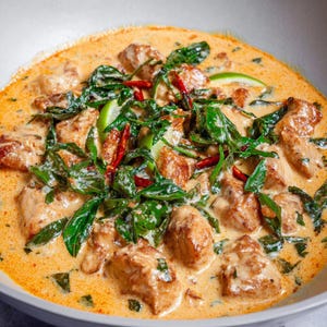 Peut inclure: Gros plan sur un plat crémeux de couleur orange avec des morceaux de viande cuite, des légumes verts à feuilles, des piments rouges et des tranches de citron vert. Le plat est servi dans un bol blanc.