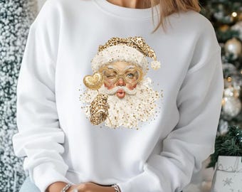 May include: White sweatshirt featuring a whimsical Santa Claus graphic. The Santa design includes a leopard print hat and glove, heart-shaped glasses, and a gold heart-shaped lollipop. The Santa is winking and blowing a kiss. A festive holiday-themed sweatshirt.