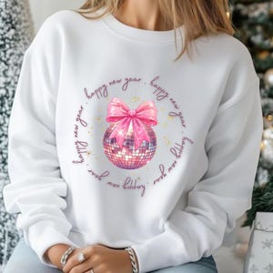 May include: White sweatshirt featuring a disco ball design with a pink bow and the words "happy new year" in a circle. The disco ball is a mix of pink and silver squares. The design includes gold star accents.