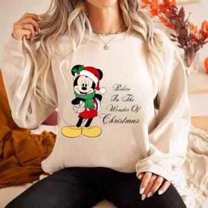 以下が含まれることがあります： サンタ帽とマフラーをつけたお祝いのミッキーマウスのグラフィックが特徴のベージュのスウェットシャツ。「Believe In The Wonder Of Christmas」の文字が右側にプリントされています。スウェットシャツは長袖で、クルーネックです。