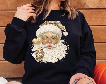 May include: Black sweatshirt featuring a whimsical Santa Claus design. Santa is depicted winking, wearing heart-shaped glasses, and a leopard print hat and glove. The design includes gold accents and a heart-shaped lollipop.