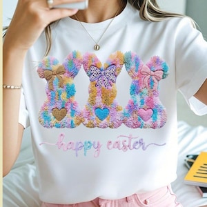 Puede incluir: Camiseta blanca con tres diseños de conejitos coloridos. Cada conejito tiene un lazo, un corazón y una textura esponjosa. Las palabras "happy easter" están bordadas en colores pastel debajo.