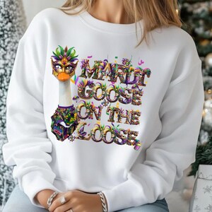 Puede incluir: Sudadera blanca con un ganso con una máscara y un collar de Mardi Gras. El texto "Mardi Goose on the Loose" está impreso en letras coloridas y festivas. El diseño incorpora tonos morados, verdes, dorados y naranjas, perfecto para un tema festivo.