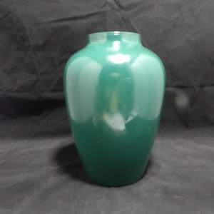 Può includere: Un vaso verde smeraldo lucido, con un corpo arrotondato e un bordo leggermente svasato. Il vaso ha una superficie liscia e riflettente, su uno sfondo scuro. Design semplice ed elegante.