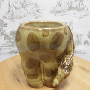 Peut inclure: Un pot en céramique en forme de girafe avec une large ouverture. Le pot est principalement vert olive avec des taches marron et une tête de girafe qui s'étend sur le côté. Le pot est posé sur une surface en bois.