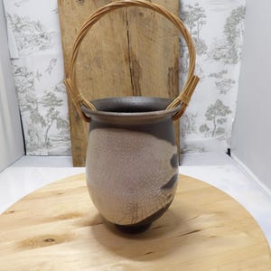May include: A ceramic vase with a crackle glaze, dark rim, and a woven handle. The vase has a bulbous shape with a dark base and a light-coloured body. The handle is made of woven, light-brown material. The vase is displayed on a wooden surface.
