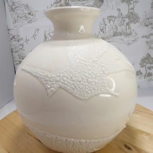 May include: Cream-colored ceramic vase with a textured, raised pattern. The vase has a rounded body and a flared neck. The textured design appears around the base and in a wave-like pattern across the body. The vase sits on a wooden surface.