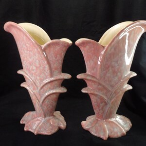 May include: Two matching vintage ceramic vases. The vases are a speckled pink with a cream-coloured interior. The vases have a tulip-like shape with a flared top and a decorative base. The vases are displayed against a black background.
