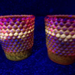 May include: Two decorative glasses with a textured, bubble-like pattern. The glasses have a color gradient, transitioning from a reddish-pink rim to yellow and brown at the base. The background is a dark blue.