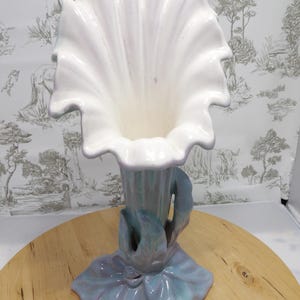 May include: A ceramic vase shaped like a large flower, with a ruffled white top and a pale lavender and blue base. The base has a leaf design. The vase is a decorative item. The vase is a decorative item, suitable for displaying flowers.
