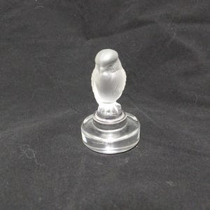 Könnte beinhalten: Eine klare Vogelfigur aus Glas mit mattierter Oberfläche. Der Vogel sitzt auf einem runden, mehrstufigen Sockel. Die Figur ist wahrscheinlich ein Dekorationsartikel oder ein Sammlerstück.