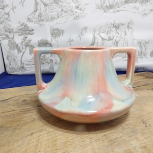 Può includere: Un vaso in ceramica con due manici caratterizzato da uno smalto sfumato nei toni pastello rosa, azzurro e crema. Il recipiente presenta una base ampia e arrotondata che si restringe verso un'apertura circolare, con una finitura liscia e lucida.
