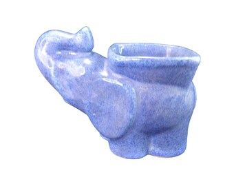 Può includere: Fioriera a forma di elefante in ceramica blu chiaro. La fioriera ha un'apertura rettangolare per le piante. La proboscide dell'elefante è sollevata e le sue orecchie sono visibili. Articolo decorativo.