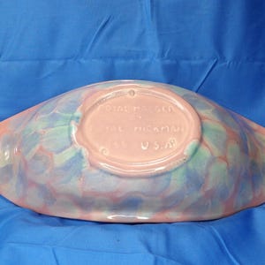 May include: A pink and blue glazed ceramic serving dish with a scalloped edge. The bottom of the dish has a pink label that reads "Gladding McBean, Tal Nickman, U.S.A."