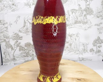 May include: A tall, maroon-colored ceramic vase with a textured surface and yellow accents. The vase has a wide, rounded top and a flared base. The yellow accents are around the top and base of the vase. The vase is on a wooden surface.