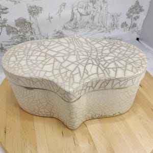May include: A decorative, off-white ceramic box with a unique, cracked glaze pattern. The box has an organic shape and sits on a round wooden surface. The lid and sides feature the intricate crackle design.