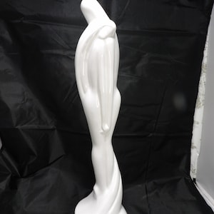 May include: A tall, white ceramic sculpture of a draped figure. The figure has a smooth, glossy finish and a flowing, elegant design. The sculpture is standing on a round base and has a stylized, abstract form.