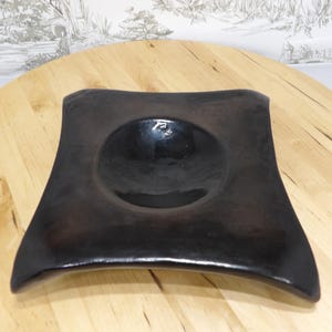 May include: A square, dark brown ceramic bowl with a rounded center. The bowl has gently curved sides and a glossy finish. The bowl is displayed on a light-colored wooden surface.