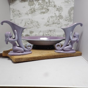 May include: A set of vintage lilac-colored ceramic vases, each featuring a kneeling woman supporting a horn-shaped vessel. A matching lilac bowl sits between the vases on a wooden cutting board. The background includes a toile wallpaper.