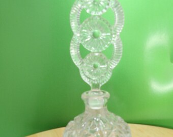 Può includere: Flacone di profumo in vetro trasparente con tappo decorativo. La bottiglia ha una base rotonda e strutturata e un tappo alto e ornato con cerchi collegati. Lo sfondo è verde.