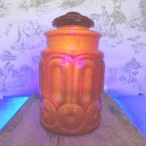 Op de afbeelding: Een oranje glazen pot met een deksel. De pot heeft een cilindrische vorm met gewelfde en cirkelvormige ontwerpen. Het deksel heeft een donkerdere oranje tint. De pot staat op een houten oppervlak.