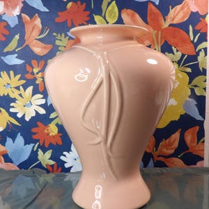 Può includere: Un vaso in ceramica color pesca con base svasata e parte superiore ampia e arrotondata. Il vaso presenta un design decorativo in rilievo di linee verticali. Lo sfondo è un motivo floreale con fiori blu, arancioni e gialli.
