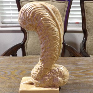 May include: A beige ceramic sculpture with a curved, feather-like design. The sculpture has a textured surface and sits on a square base. The piece is displayed on a wooden table, showcasing its artistic form.