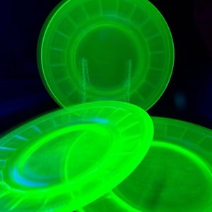 Könnte beinhalten: Drei leuchtend grüne Glasplatten. Die Platten haben ein kreisförmiges Design mit erhabenem Rand und einem zentralen, vertieften Bereich. Die Platten sind in einer dynamischen Komposition vor dunklem Hintergrund angeordnet, was ihre lebendige Farbe hervorhebt.