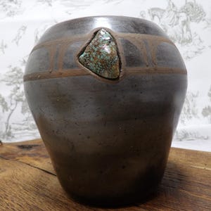 May include: A dark brown ceramic vase with a rounded body and a wide opening. The vase features a decorative band and a triangular turquoise stone inlay. The vase is displayed on a wooden surface.