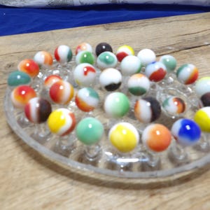 Puede incluir: Un tablero ovalado de vidrio transparente con múltiples hendiduras circulares, cada una con una pequeña canica de vidrio multicolor. Las canicas presentan varios patrones opacos y translúcidos en tonos rojo, naranja, amarillo, verde, azul, blanco y negro.