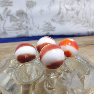 May include: Four spherical glass marbles featuring a white base with swirling bands of reddish-orange color. The marbles are resting in the circular indentations of a clear, molded glass flower frog or display stand with multiple openings.