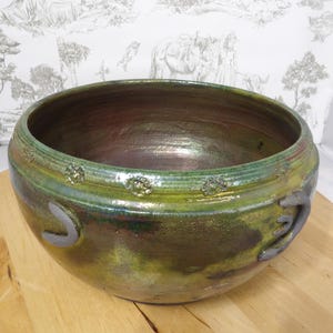 May include: A handcrafted ceramic bowl with a wide, open design. The bowl features a glossy finish with shades of green, brown, and gold. Decorative elements include raised floral patterns and gray, curved accents. The bowl sits on a wooden surface.