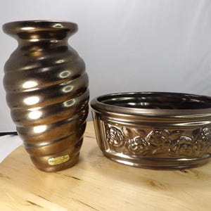 Può includere: Un set di vasi e fioriere in ceramica color bronzo. Il vaso ha un design a spirale e la fioriera presenta un motivo floreale. Entrambi i pezzi hanno una finitura lucida e sono esposti su una superficie di legno.