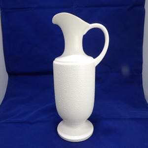 May include: A white ceramic jug with a textured surface. The jug has a handle and a spout, and it sits on a round base. The jug is approximately 30 cm tall.