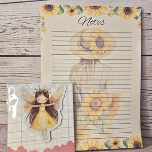 Könnte beinhalten: Ein Notizblock und ein magnetischer Lesezeichen mit Aquarellillustrationen. Der Notizblock hat ein Sonnenblumenmuster und das Wort "Notizen". Das Lesezeichen zeigt eine Fee in einem gelben Kleid und Flügeln.