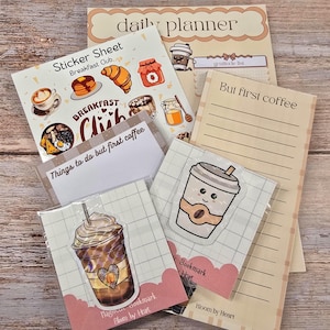 Può includere: Una collezione di articoli di cancelleria a tema caffè, tra cui un "Daily Planner", un "Sticker Sheet" con illustrazioni della colazione e segnalibri. Gli articoli presentano illustrazioni di tazze di caffè e la frase "But first coffee".