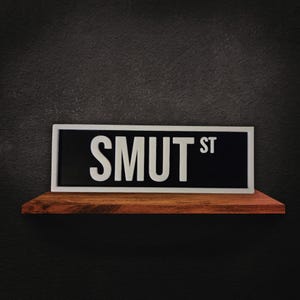 Puede incluir: Un letrero de calle rectangular con el texto "SMUT ST" en letras blancas sobre un fondo negro, enmarcado por un borde blanco. El letrero descansa sobre una estanter&iacute;a de madera contra una pared gris oscura.