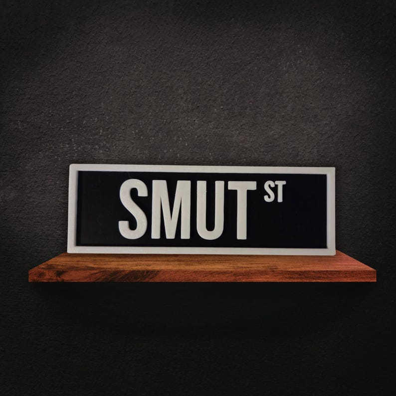 Puede incluir: Un letrero de calle rectangular con el texto "SMUT ST" en letras blancas sobre un fondo negro, enmarcado por un borde blanco. El letrero descansa sobre una estanter&iacute;a de madera contra una pared gris oscura.