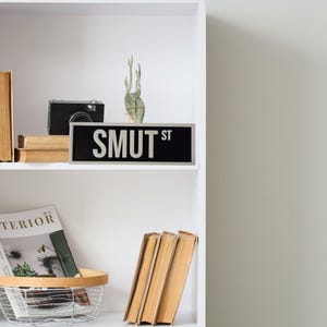 Puede incluir: Una estanter&iacute;a blanca exhibe un letrero negro que dice "SMUT ST" en letras blancas. Una c&aacute;mara negra, libros peque&ntilde;os y un cactus tambi&eacute;n est&aacute;n en el estante. Una revista y libros est&aacute;n en el estante inferior.